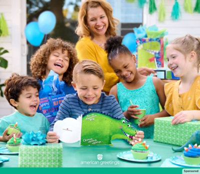 A young boy reads a dinosaur birthday card, surrounded by smiling family and friends.