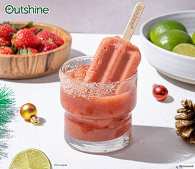 Outshine bar submerged in a cocktail glass on a table with holiday decor and bowls of fresh fruit.