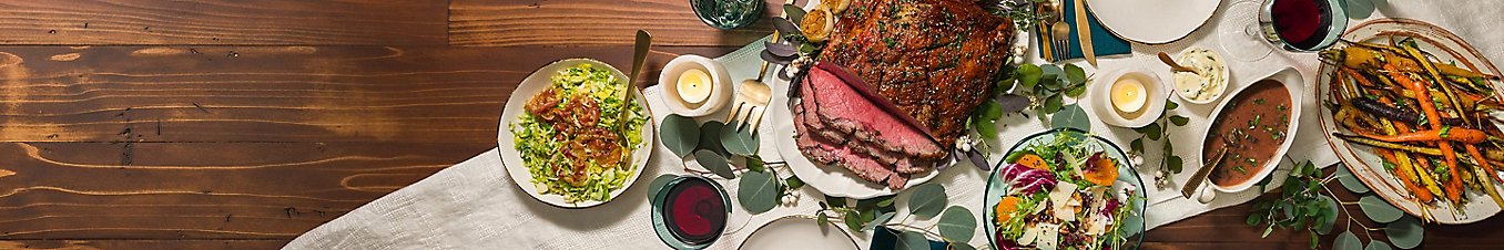 A holiday dinner with salads and roast beef with a side of sauce.