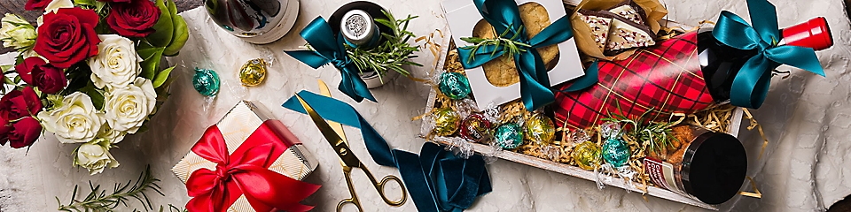 A holiday gift box with cookies, chocolates, wine, and almonds placed next to some roses and other wrapped gifts.