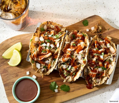 Three pork carnita tacos on wooden cutting board with lime wedges and dipping sauce.