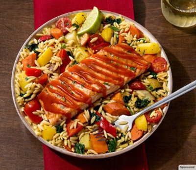 Bowl of orzo, vegetables and salmon on a wooden table with a red, cloth napkin.