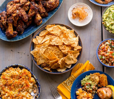 Table assortment of various foods such as chicken wings, chips, macaroni, salsas, and shrimp.