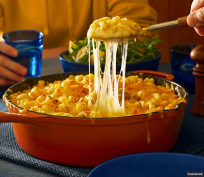 Melty mac & cheese being spooned out of a deep dish pan at the dinner table.