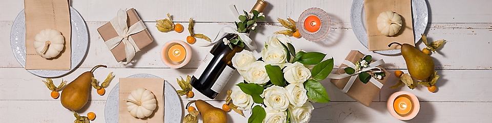 A bottle of wine, a bouquet of white roses, plates, candles and pumpkin decors laid out on the floor.